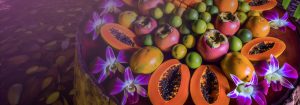 whole fresh fruits arranged in a circular array in a natural tropical setting. The fruits include persimmon, lime, orange, and mango, with sliced papayas showing their seeds interspersed throughout.